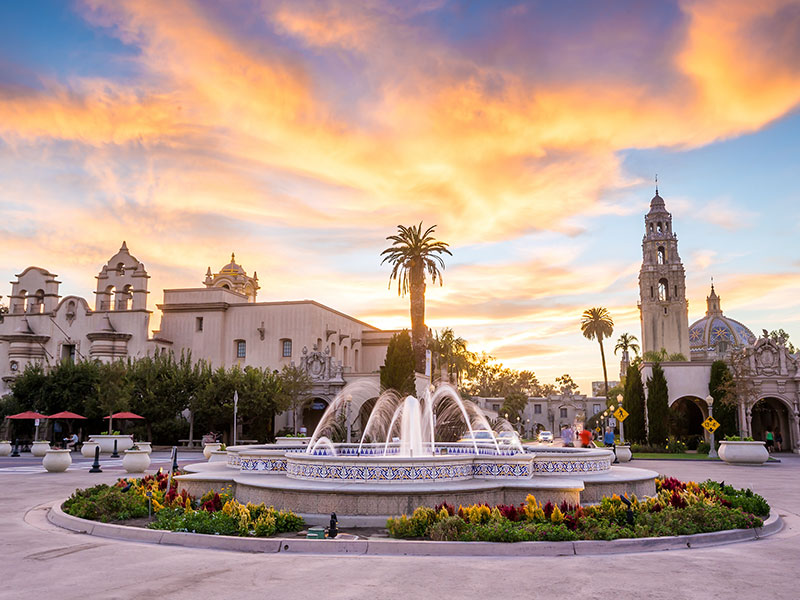 A large fountain in front of a building with a palm tree and colorful clouds in the sky.